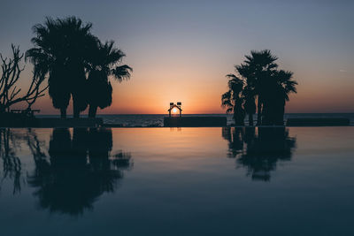 Silhouette palm trees by swimming pool against sky during sunset