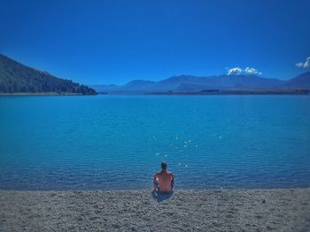 Rear view of shirtless man sitting at beach against blue sky