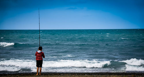 Rear view of boy fishing at beach against blue sky