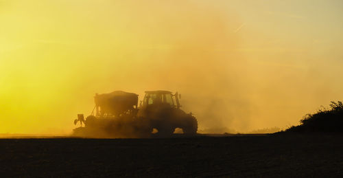 Silhouette of a tractor sowing seeds in a field in a cloud of dust against the background.