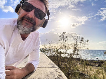 Portrait of young man wearing sunglasses while sitting at beach against sky during sunset