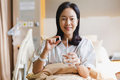 Woman holding white pills and a glass of water while sitting on a bed in hospital