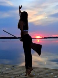 Playful woman holding broom while jumping on pier by lake during sunset