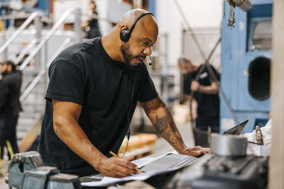 Mature male factory worker talking through headset while using laptop in warehouse