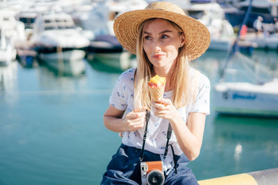 Smiling woman wearing hat eating ice cream sitting at harbor