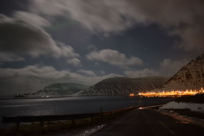 Road by illuminated mountains against cloudy sky at night