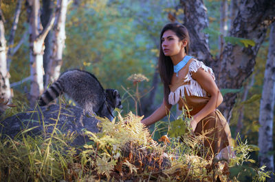 Young woman in traditional clothing standing by raccoon in forest