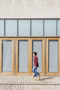 Full body side view of active young male in checkered shirt and jeans performing trick on unicycle near mirrored glass wall of contemporary building