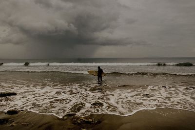 Man carrying surfboard while standing on beach against cloudy sky