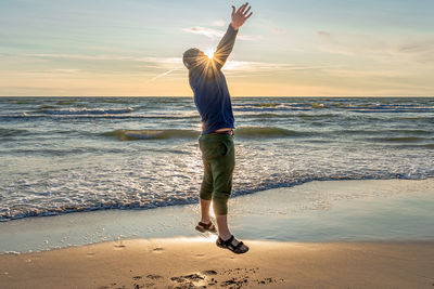 Full length of man jumping with arms raised at beach during sunset