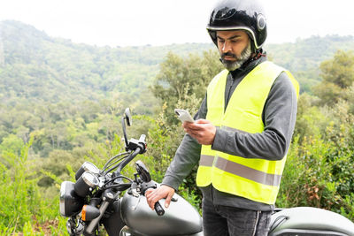 Thoughtful adult hispanic male biker in protective helmet and vest messaging on mobile phone while standing near broken motorbike near lush green woods