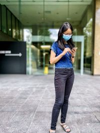 Portrait of asian woman in eyeglasses and surgical face mask using smartphone against building.