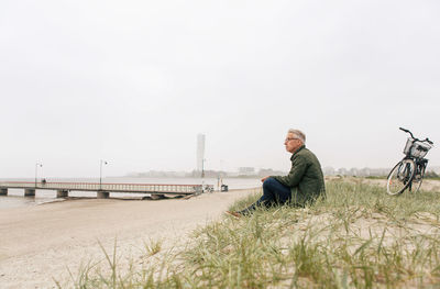 Full length of senior male commuter sitting on sand at beach against clear sky