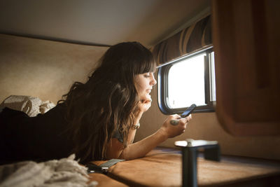 Woman relaxing on bed by window while using smart phone in camper van