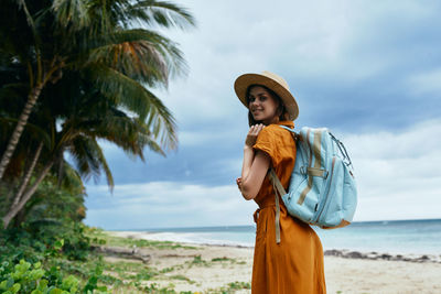 Young woman standing by palm tree on beach against sky