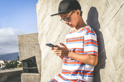Boy wearing cap using smart phone while standing against wall outdoors