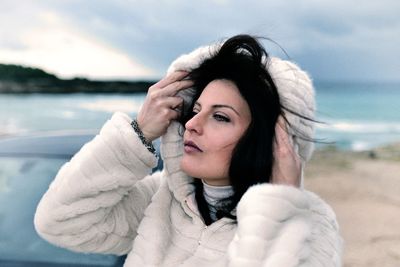 Young beautiful woman wearing winter coat at beach against sky