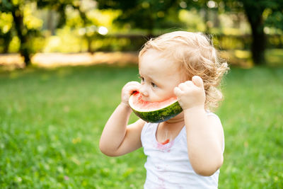 Funny smiling kid boy in white bodysuit eating watermelon at green lawn