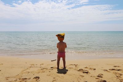 Rear view of shirtless baby boy standing at beach against cloudy sky