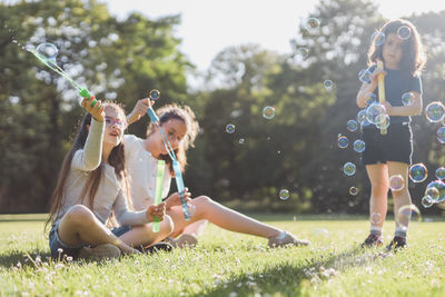 Three caucasian girls are sitting on a flower lawn in a public park and blowing soap bubbles