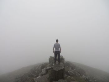 Rear view of man standing on rock against sky during foggy weather