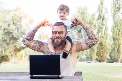 Smiling bearded man using laptop while son sitting on shoulder in park