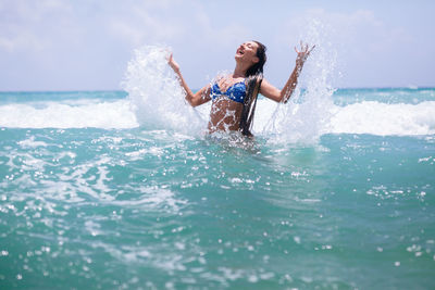 Full length of young woman splashing water in sea against sky