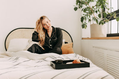 A happy pretty young woman in pajamas is resting and having breakfast in bed in a cozy apartment