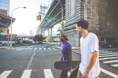 Young man with woman walking on road while holding skateboard