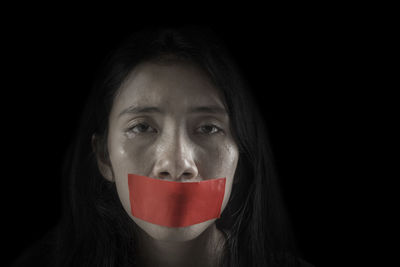 Close-up portrait of crying young woman with covered mouth with tape against black background