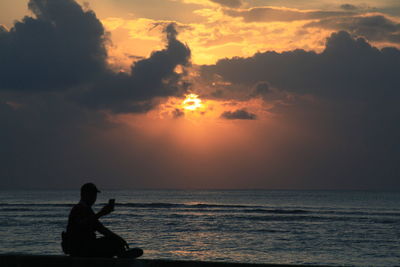 Silhouette man sitting by sea against sky during sunset