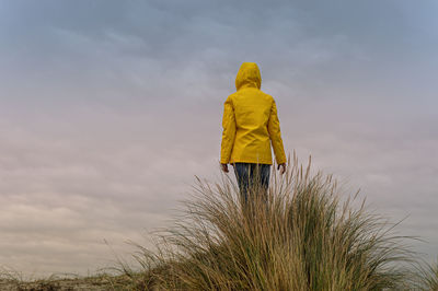 Woman wearing a yellow coat standing on top of a dune, rear view, book cover concept.