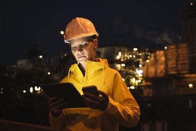 Female professional in yellow hardhat and raincoat using wireless technologies at night