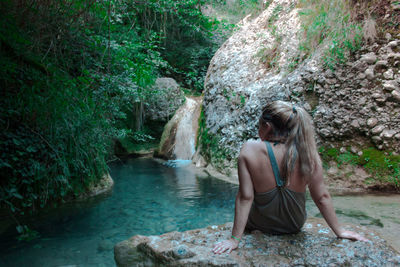 Rear view of woman sitting on rock by pond in forest