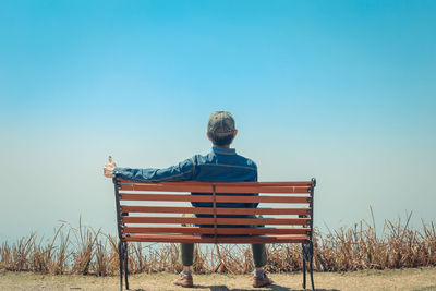 Rear view of man sitting on bench against blue sky