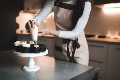 Woman decorate chocolate cupcakes with whipped cream in pastry bag closeup in kitchen.