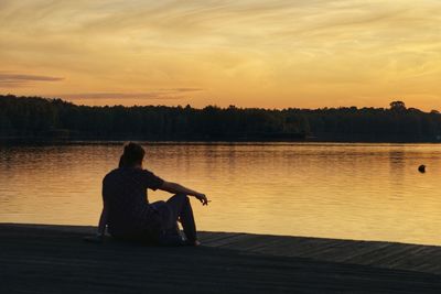 Silhouette man sitting by lake against sky during sunset