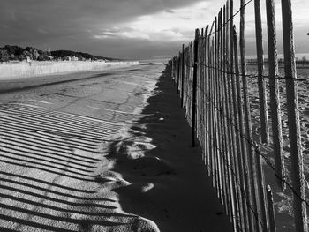 Shadow of fence falling on sand at beach against cloudy sky