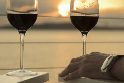 Cropped hand of man holding wine glass by sea against sky during sunset