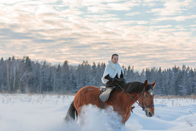 A girl in a white cloak rides a brown horse in winter. golden hour, setting sun.