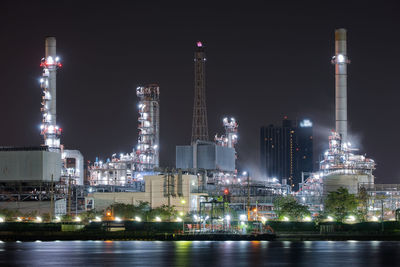 Illuminated factory by river against sky at night