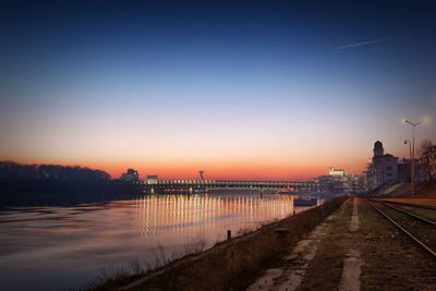 Illuminated city by river against sky at sunset