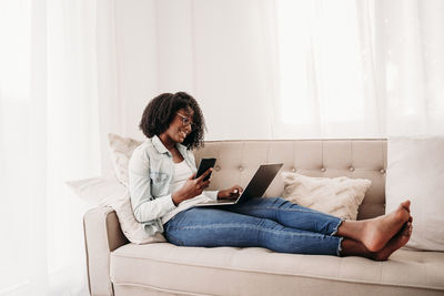 Young freelancer with smart phone and laptop sitting on sofa at home