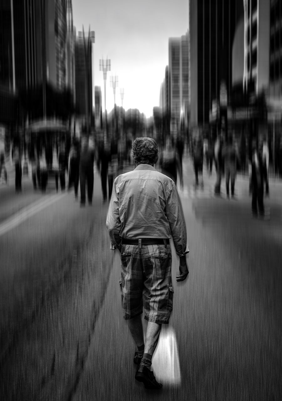 REAR VIEW OF MAN WALKING ON STREET AGAINST CITY