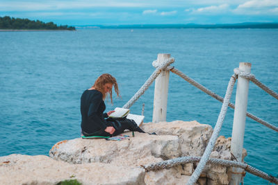 Rear view of woman sitting on rock by sea against sky