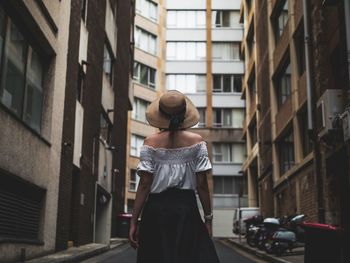 Rear view of woman wearing hat standing on road amidst buildings