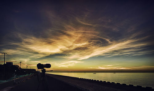 Silhouette camera on road by sea against cloudy sky during sunset