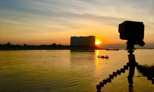 Silhouette buildings by river against sky during sunset