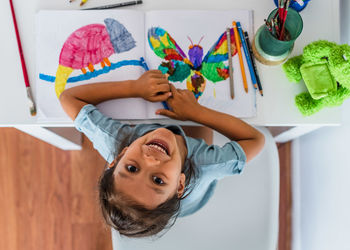 High view of a little girl sitting on the table and painting pictures of animals.