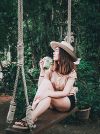 Woman wearing hat sitting by plants against trees